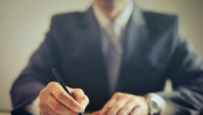 Vīrietis uzvalkā sēž pie galda, labajā rokā pildspalva