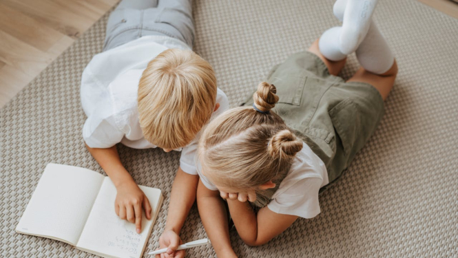 Divi bērni, zēns un meitene, guļ uz grīdas un skatās grāmatā