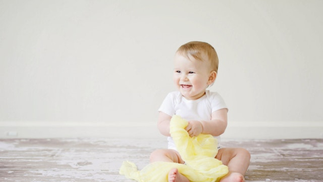 Smaidošs mazulis sēž uz grīdas, turpt rokā dzeltenu lakatu