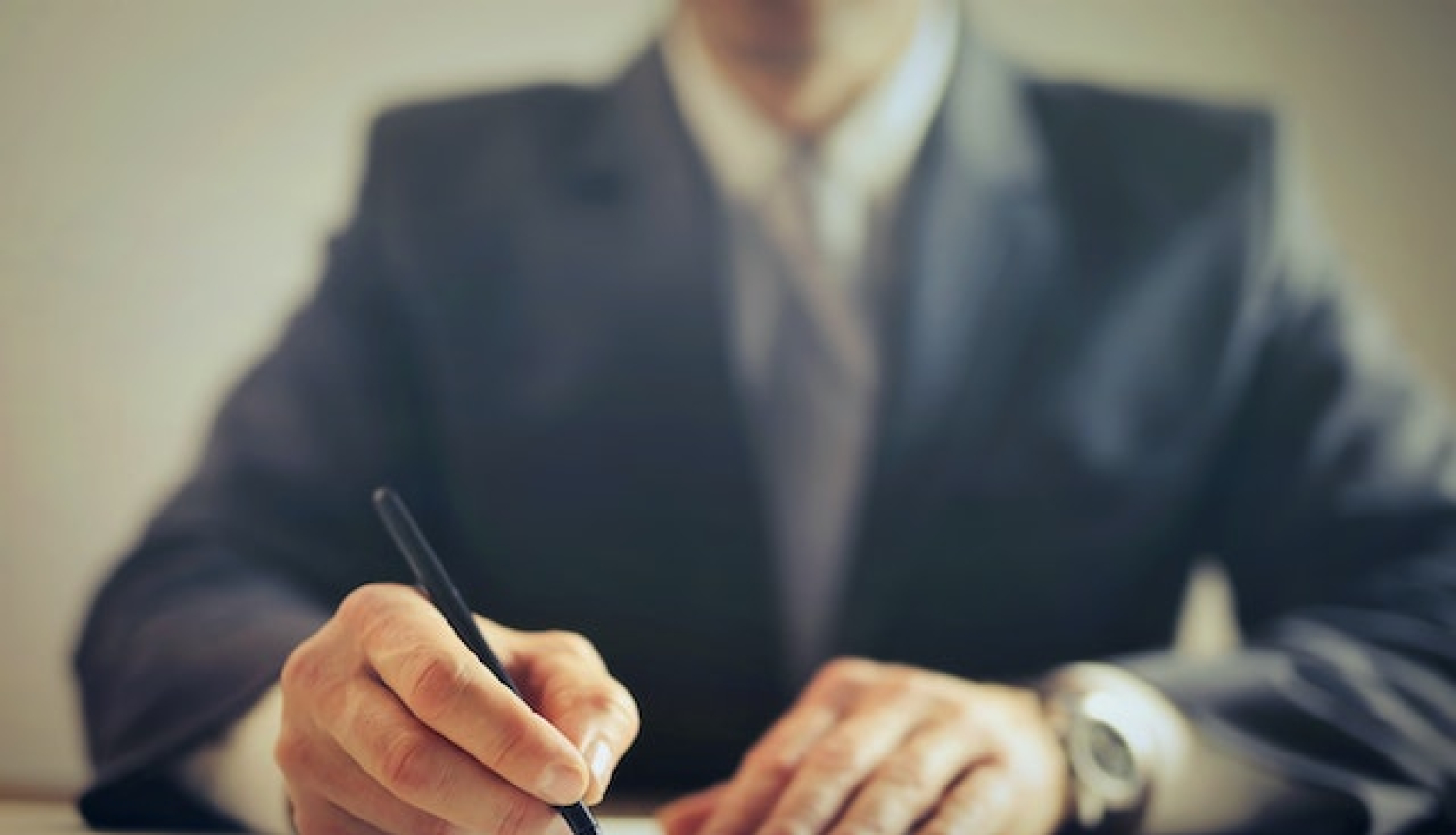 Vīrietis uzvalkā sēž pie galda, labajā rokā pildspalva