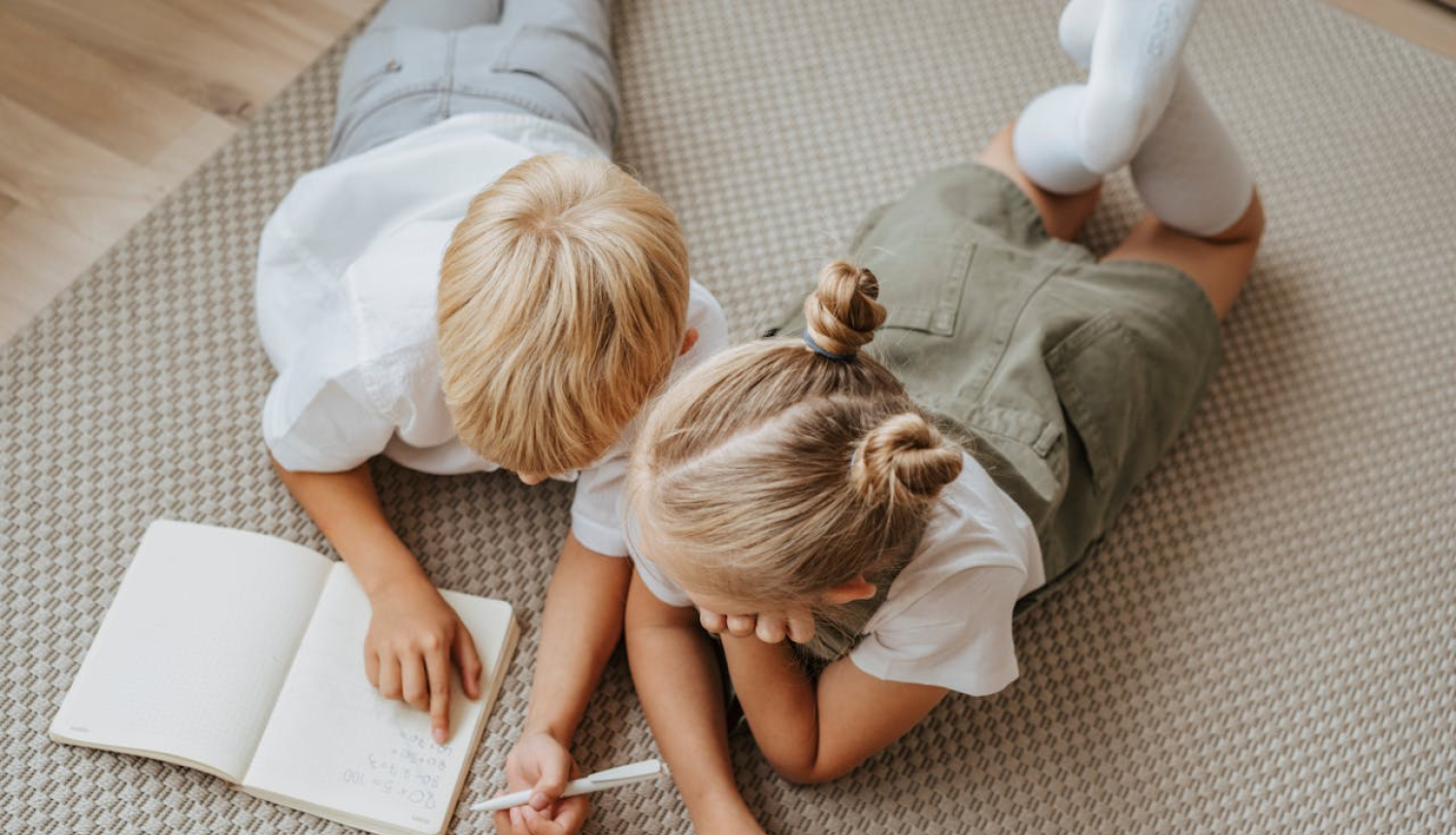 Divi bērni, zēns un meitene, guļ uz grīdas un skatās grāmatā