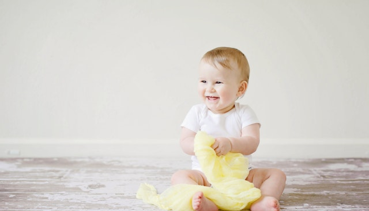 Smaidošs mazulis sēž uz grīdas, turpt rokā dzeltenu lakatu